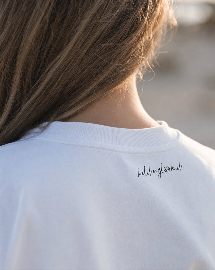 Schwarzer Schriftzug „heldenglück.de“ außen im Nacken des weißen T-Shirts, direkt unter dem Kragen.
