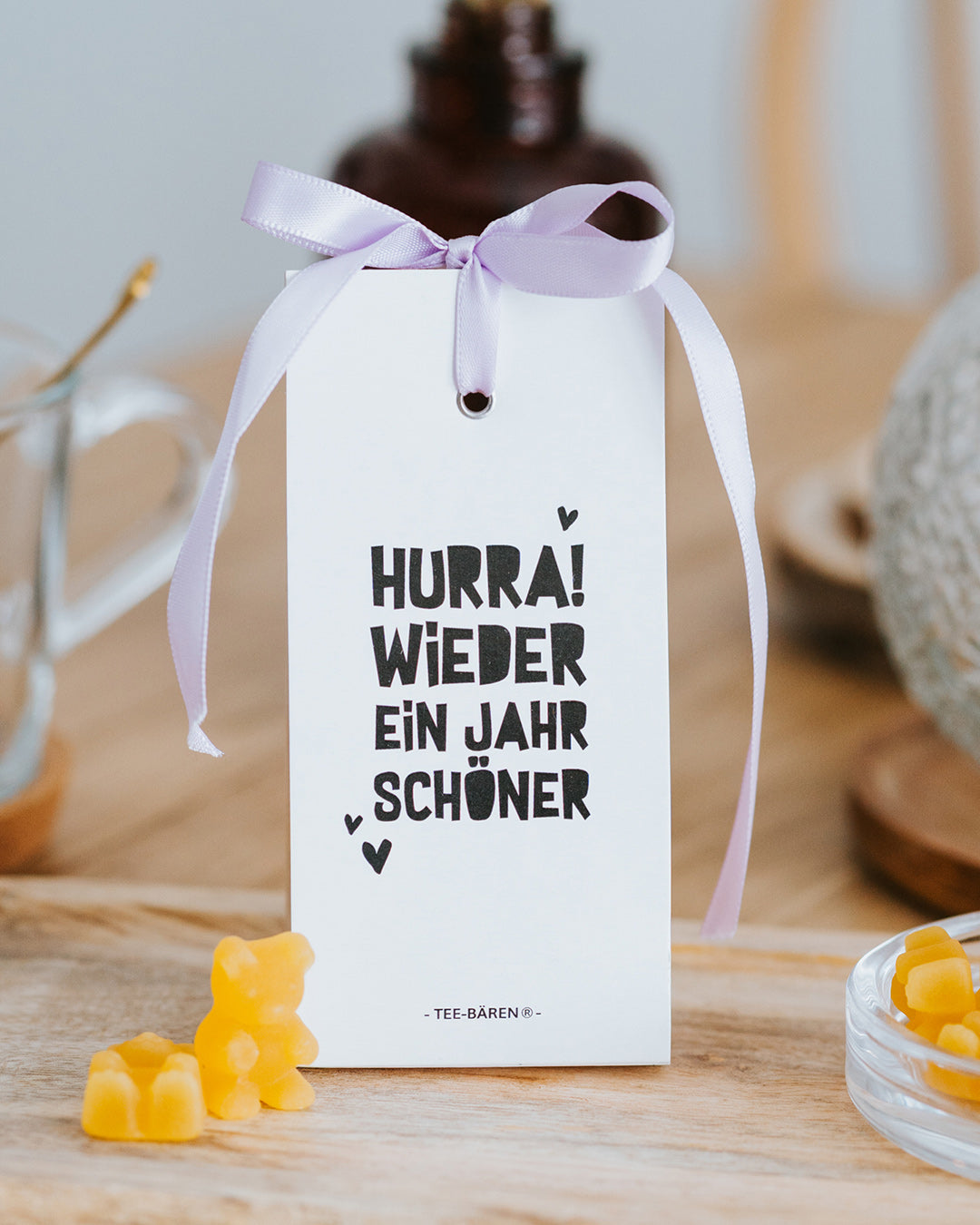 Teebären Hurra! Wieder ein Jahr schöner mit fliederfarbener Schleife, gelbe Bärchen davor.