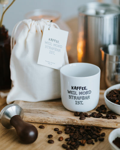 Porzellanbecher „Kaffee, weil Mord strafbar ist.“ neben Baumwollbeutel mit Anhänger und Kaffeebohnen.