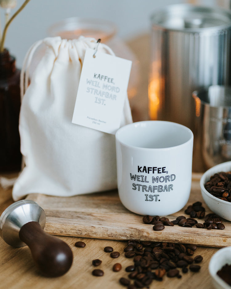 Porzellanbecher „Kaffee, weil Mord strafbar ist.“ neben Baumwollbeutel mit Anhänger und Kaffeebohnen.