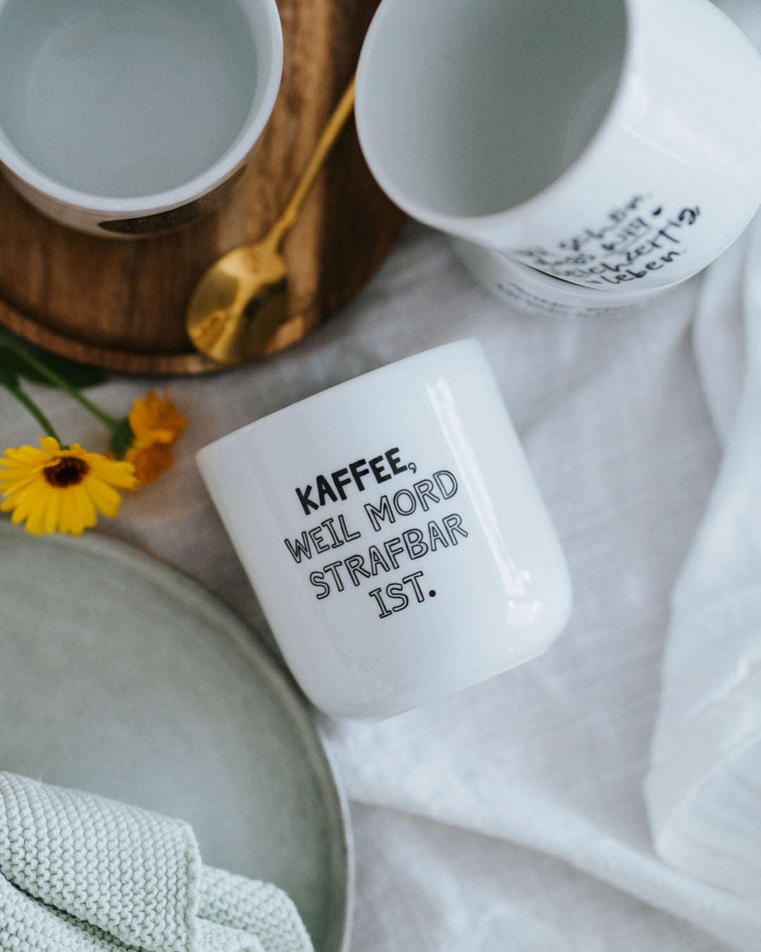 Porzellanbecher „Kaffee, weil Mord strafbar ist.“ liegend auf hellem Tuch – minimalistisches, zeitloses Design.