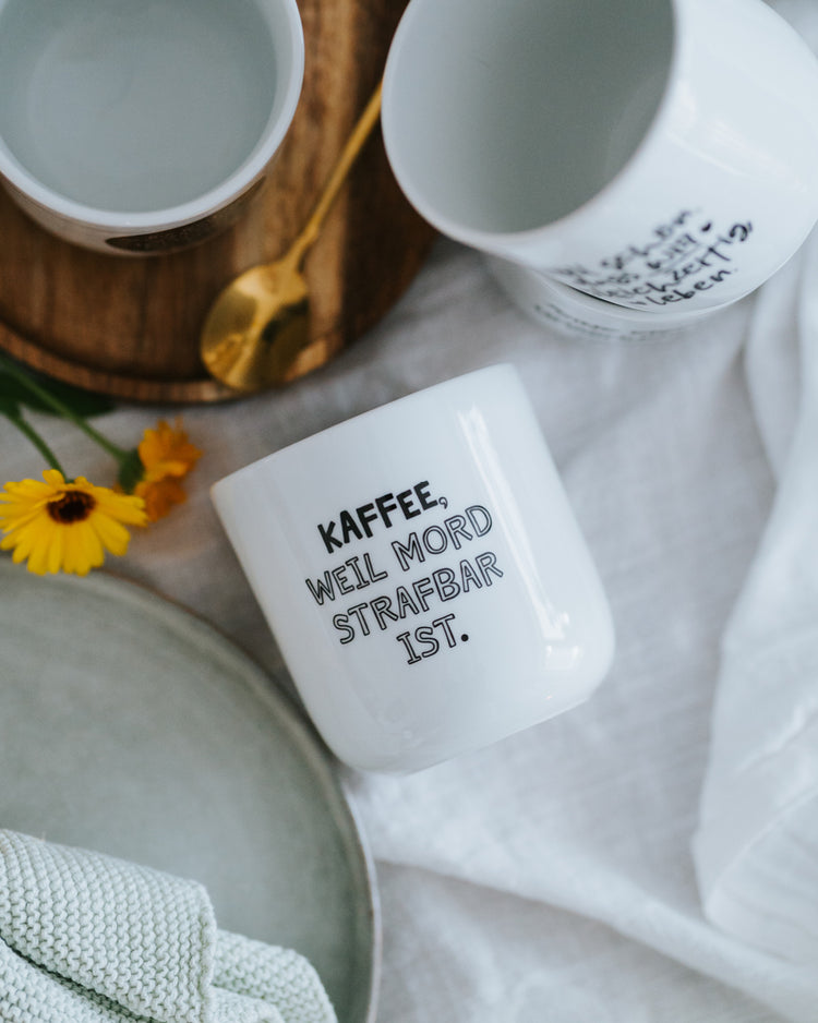 Porzellanbecher „Kaffee, weil Mord strafbar ist.“ liegend auf hellem Tuch – minimalistisches, zeitloses Design.