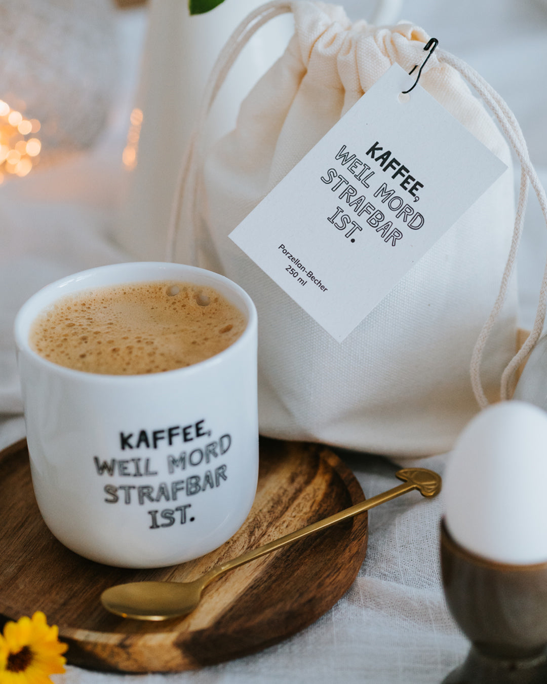 Porzellanbecher „Kaffee, weil Mord strafbar ist.“ mit Milchkaffee, dahinter Geschenkbeutel und Ei.