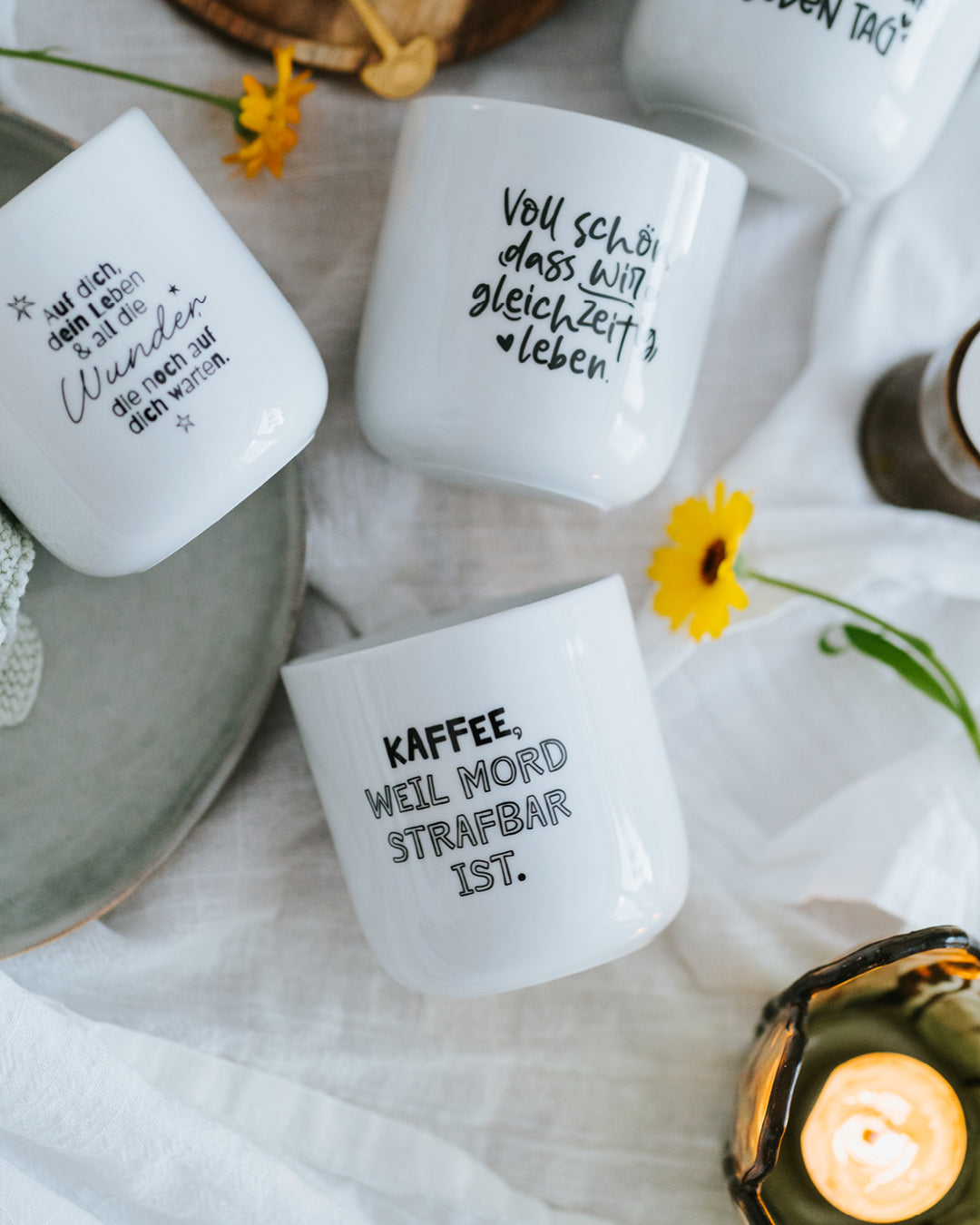 Porzellanbecher „Kaffee, weil Mord strafbar ist.“ neben weiteren Motiven und Windlicht auf weißem Tuch.