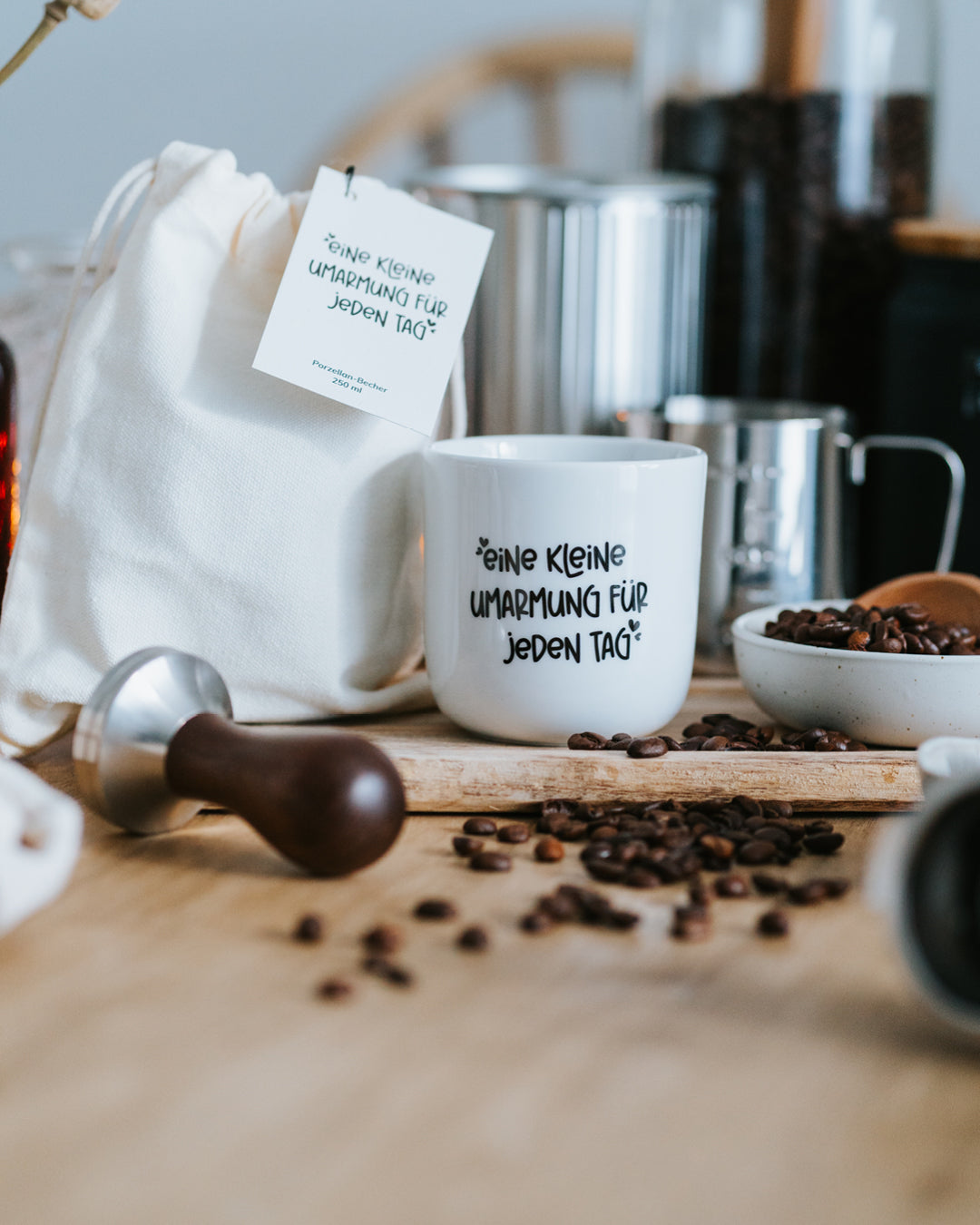 Porzellanbecher „Kleine Umarmung“ neben Baumwollbeutel, Milchkännchen und Kaffeebohnen.
