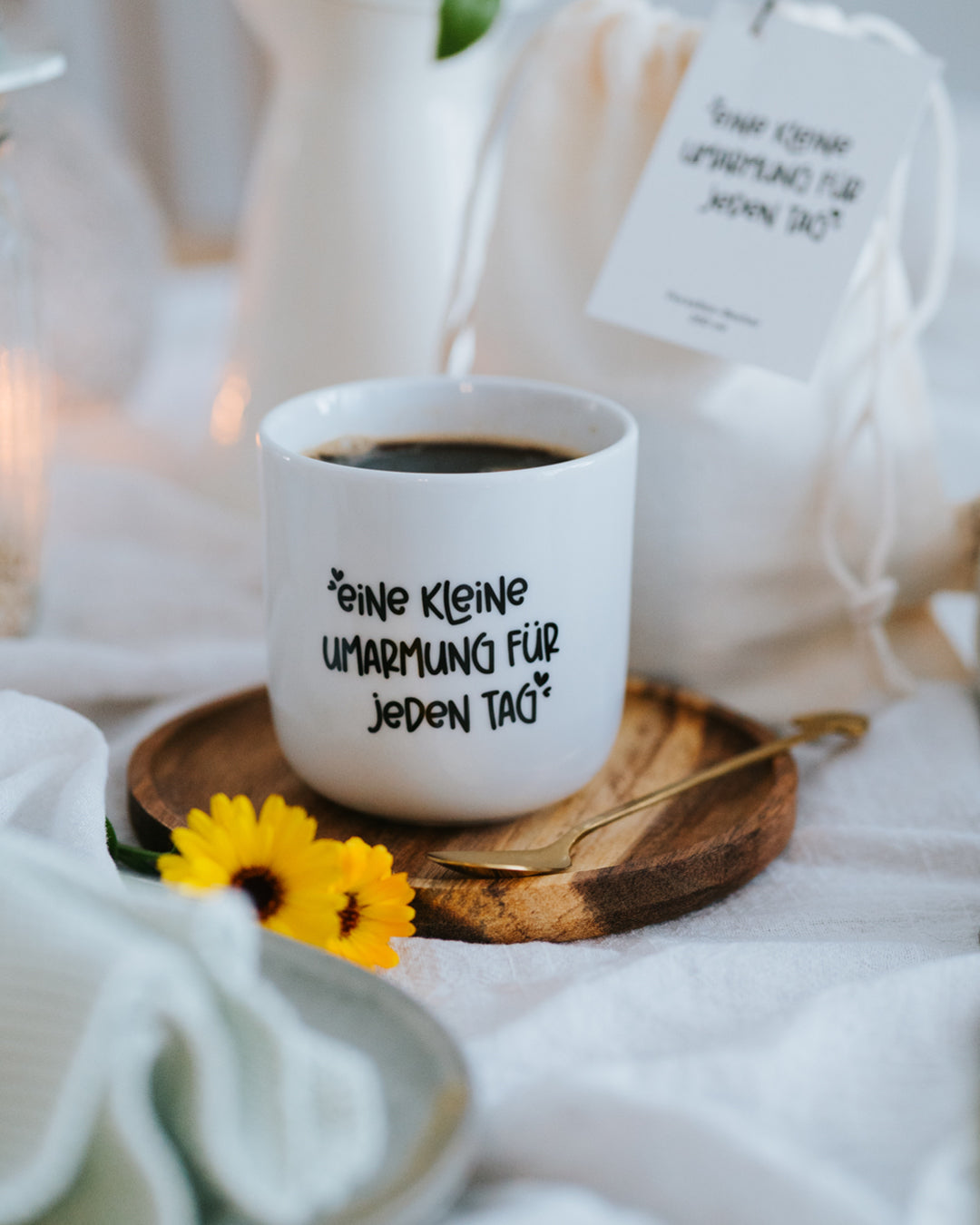 Porzellanbecher „Kleine Umarmung“ mit Kaffee auf Holzuntersetzer, Geschenkanhänger im Hintergrund.