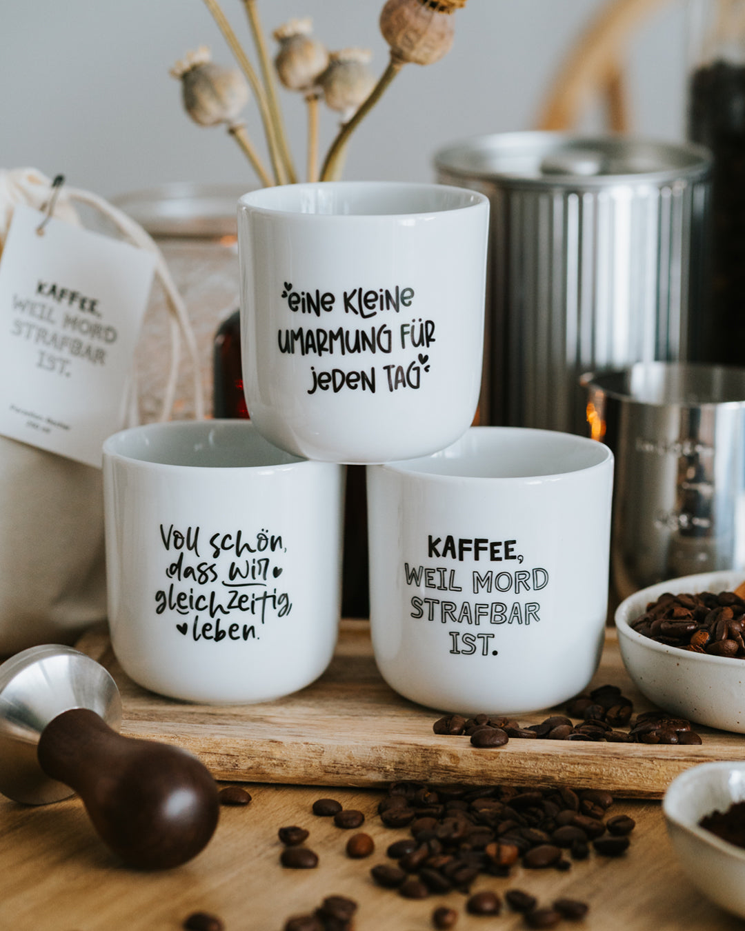 Drei Porzellanbecher nebeneinander, „Kleine Umarmung“ mittig, dazu Tamper und Kaffeebohnen.