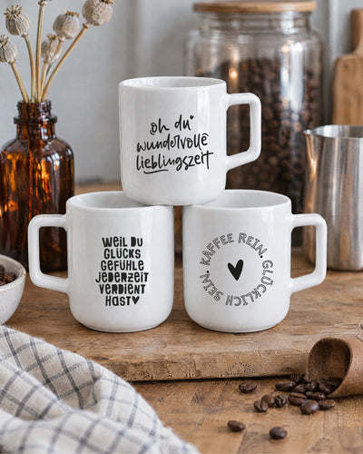 Drei Porzellantassen mit verschiedenen Sprüchen vor Kaffeebohnen und Milchkännchen.