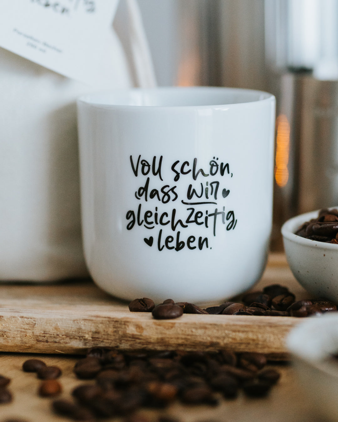 Porzellanbecher „Voll schön, dass wir gleichzeitig leben“ auf Holzbrett mit Kaffeebohnen.