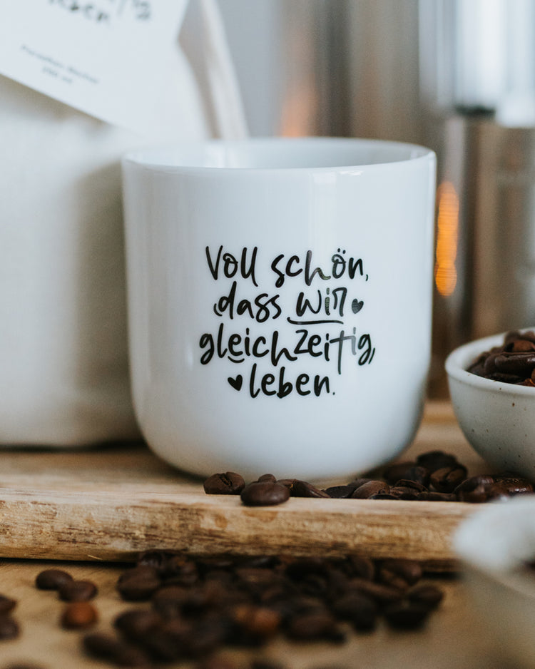 Porzellanbecher „Voll schön, dass wir gleichzeitig leben“ auf Holzbrett mit Kaffeebohnen.