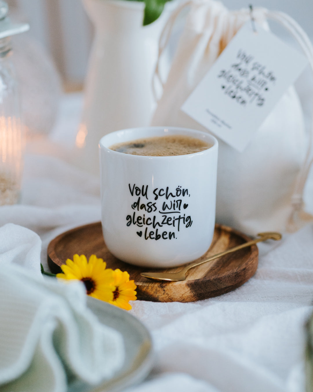 Porzellanbecher „Voll schön, dass wir gleichzeitig leben“ mit Milchkaffee auf Holzteller.