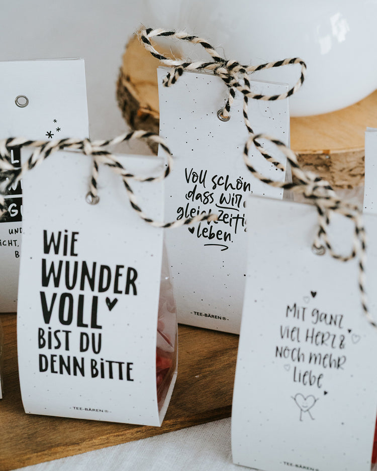 Drei Teebären-Motive mit schwarz-weißer Kordel nebeneinander, dahinter eine Teekanne.