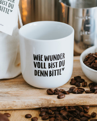 Porzellanbecher „Wie wundervoll bist du denn bitte“ auf Holzbrett mit Kaffeebohnen – zeitloses Design.
