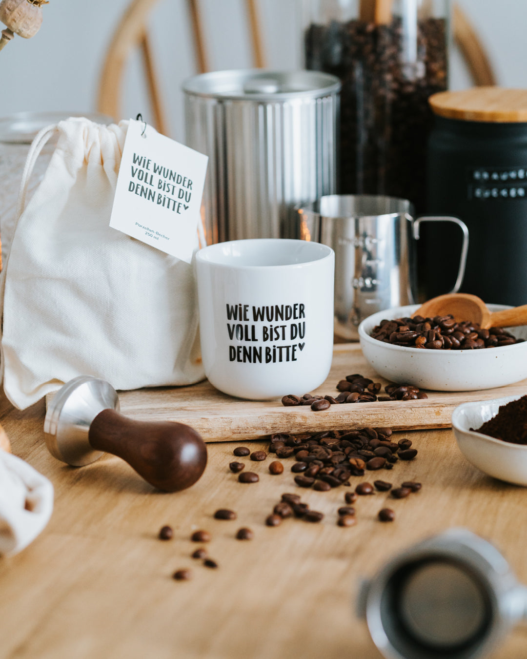 Porzellanbecher „Wie wundervoll“ neben Baumwollbeutel mit Anhänger, Milchkännchen und Kaffeebohnen.