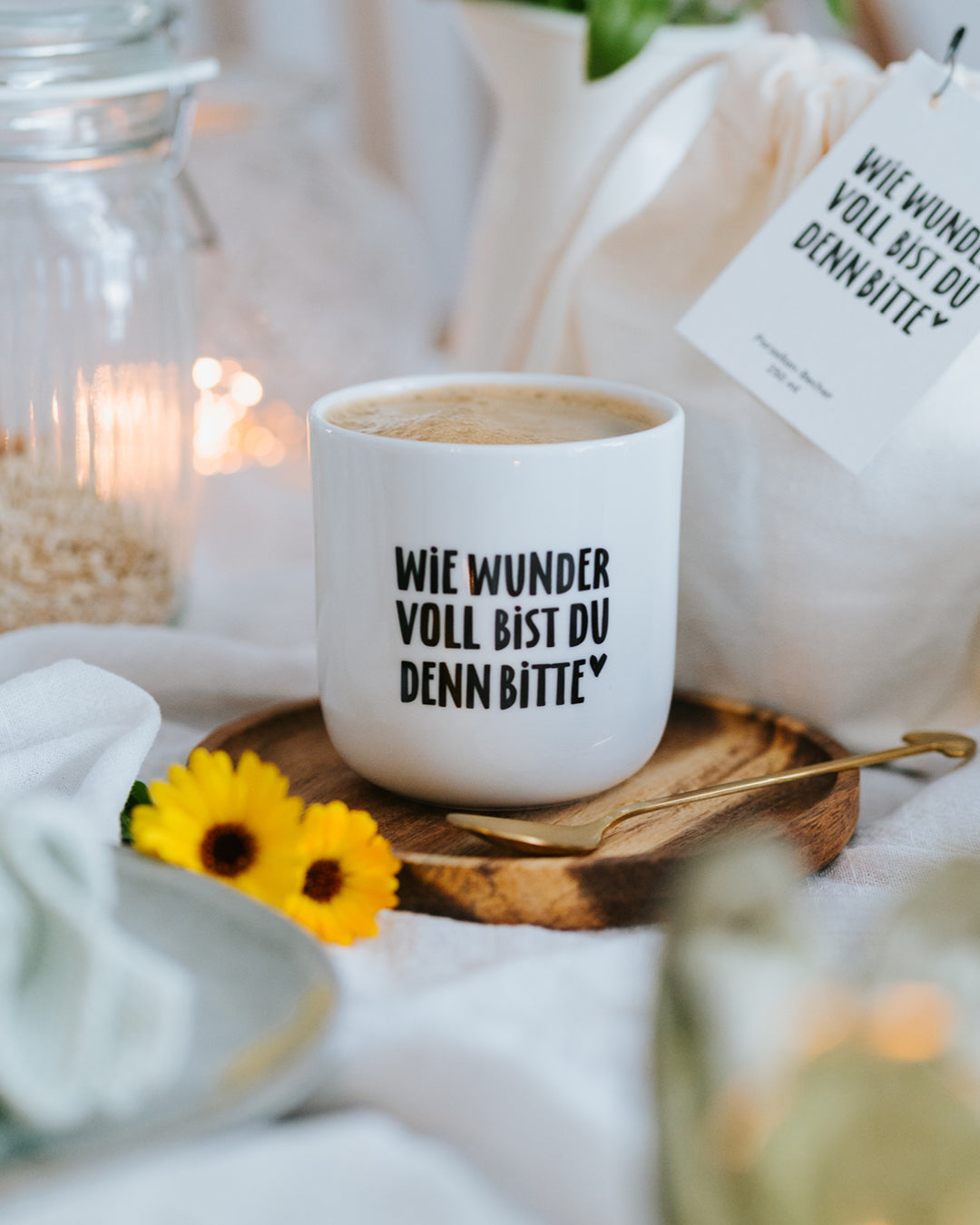 Porzellanbecher „Wie wundervoll“ mit Milchkaffee auf Holzteller mit Blume und Goldlöffel.