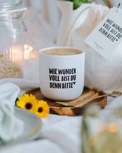 Porzellanbecher „Wie wundervoll“ mit Milchkaffee auf Holzteller mit Blume und Goldlöffel.