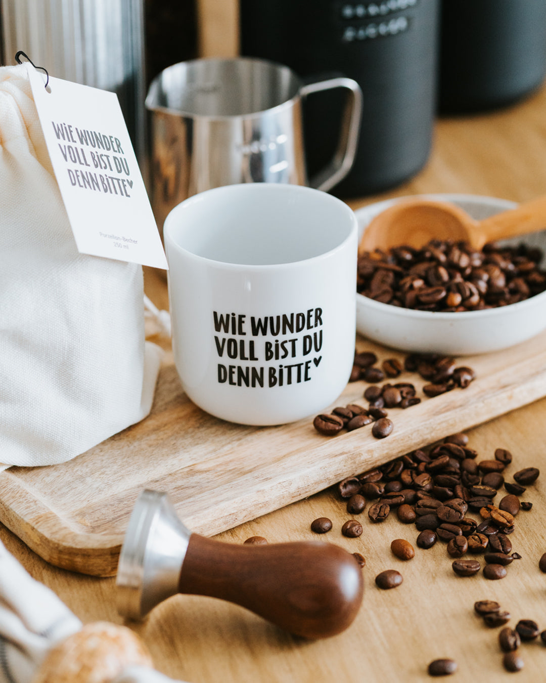 Porzellanbecher „Wie wundervoll“ auf Holzbrett mit Milchkännchen, Tamper und Kaffeebohnen.