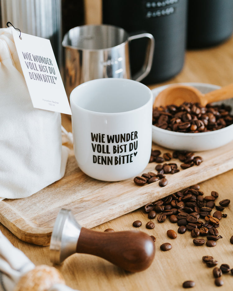 Porzellanbecher „Wie wundervoll“ auf Holzbrett mit Milchkännchen, Tamper und Kaffeebohnen.
