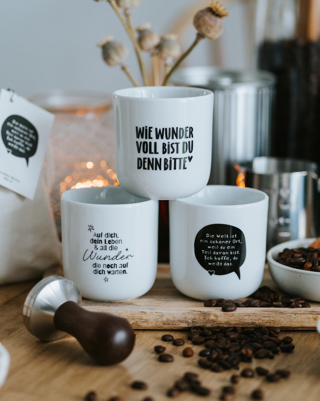 Drei Porzellanbecher auf Holzbrett, „Wie wundervoll“ hinten, Tamper und Kaffeebohnen davor.