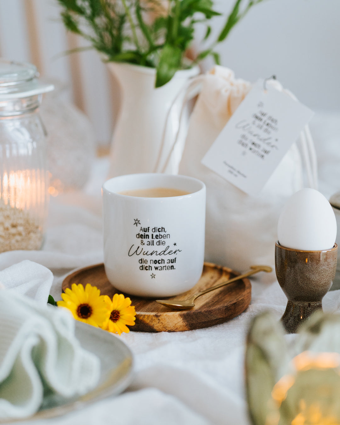 Porzellanbecher „Wunder“ mit Kaffee auf gedecktem Frühstückstisch mit Blumen und Ei im Eierbecher.