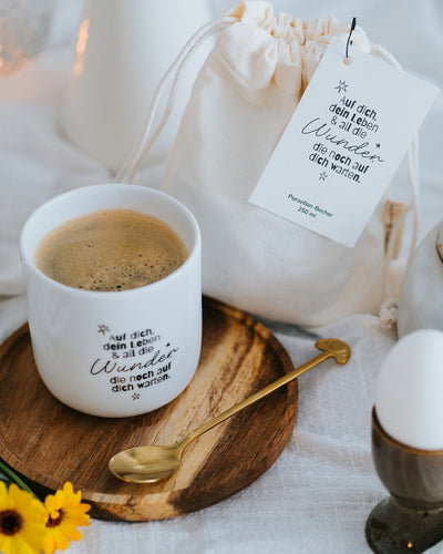 Porzellanbecher „Wunder“ gefüllt mit Kaffee auf Holzteller mit Goldlöffel – ideal für Kaffee oder Tee.