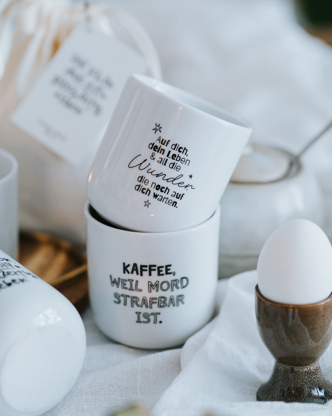 Porzellanbecher „Wunder“ und „Kaffee, weil Mord strafbar ist.“ nebeneinander auf weißem Tuch.