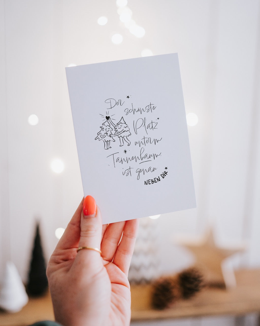 Hand hält die Postkarte „Der schönste Platz unterm Tannenbaum“ vor unscharfen Lichterketten im Hintergrund