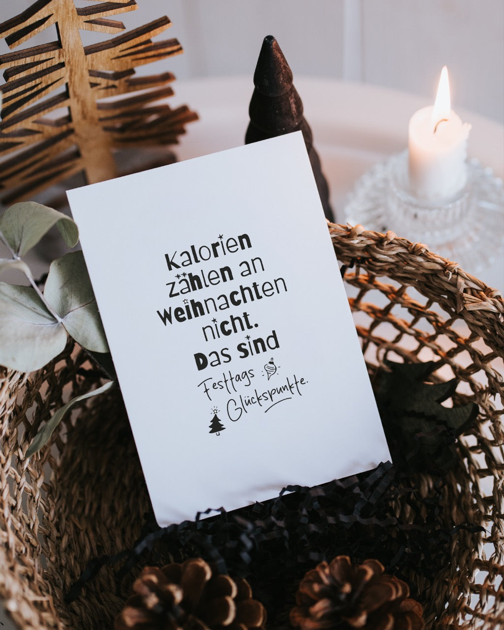 Weihnachtspostkarte „Festtags-Glückspunkte“ steht in einem Korb zwischen Eukalyptuszweig, dunkler Tannenbaum-Figur und brennender Kerze