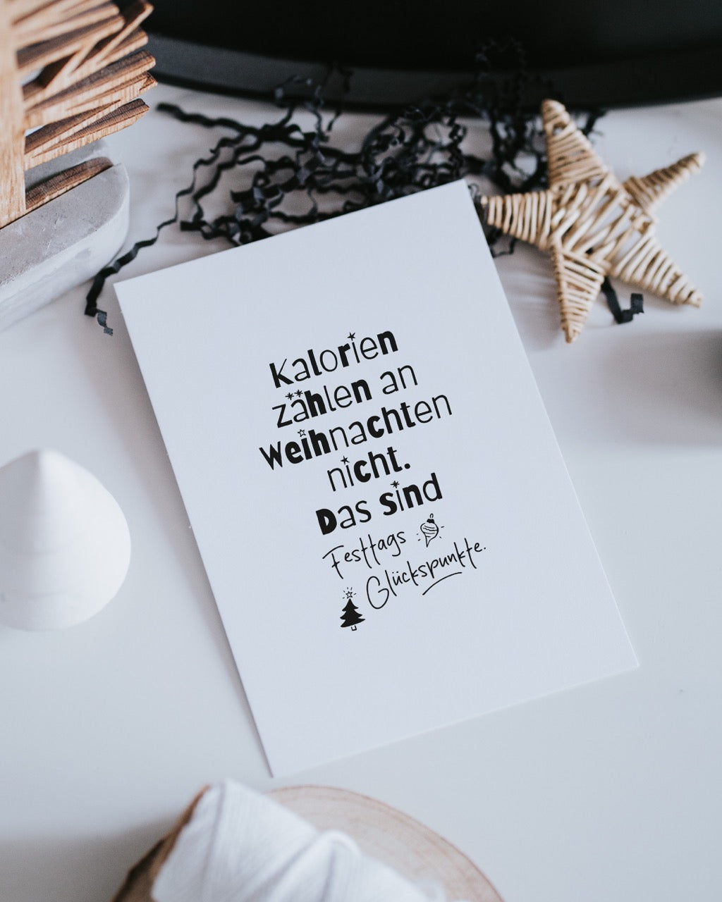 Postkarte „Festtags-Glückspunkte“ liegt auf heller Fläche neben Holz-Weihnachtsbaum und geflochtenem Stern