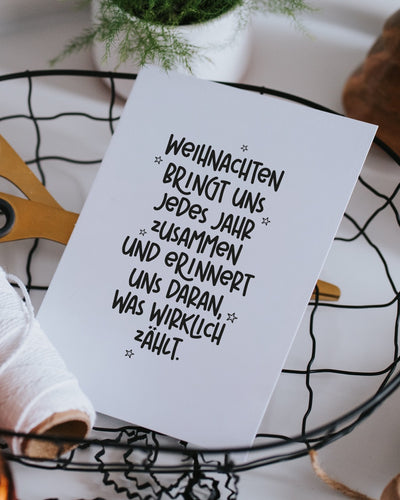 Postkarte „Was wirklich zählt“ mit Weihnachtsspruch in einem Drahtkorb neben goldener Schere und brennender Kerze