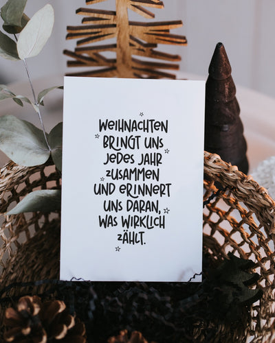 Postkarte „Weihnachten bringt uns jedes Jahr zusammen“ steht zwischen Eukalyptuszweig, Holz-Christbaum und dunkler Tannenbaum-Figur