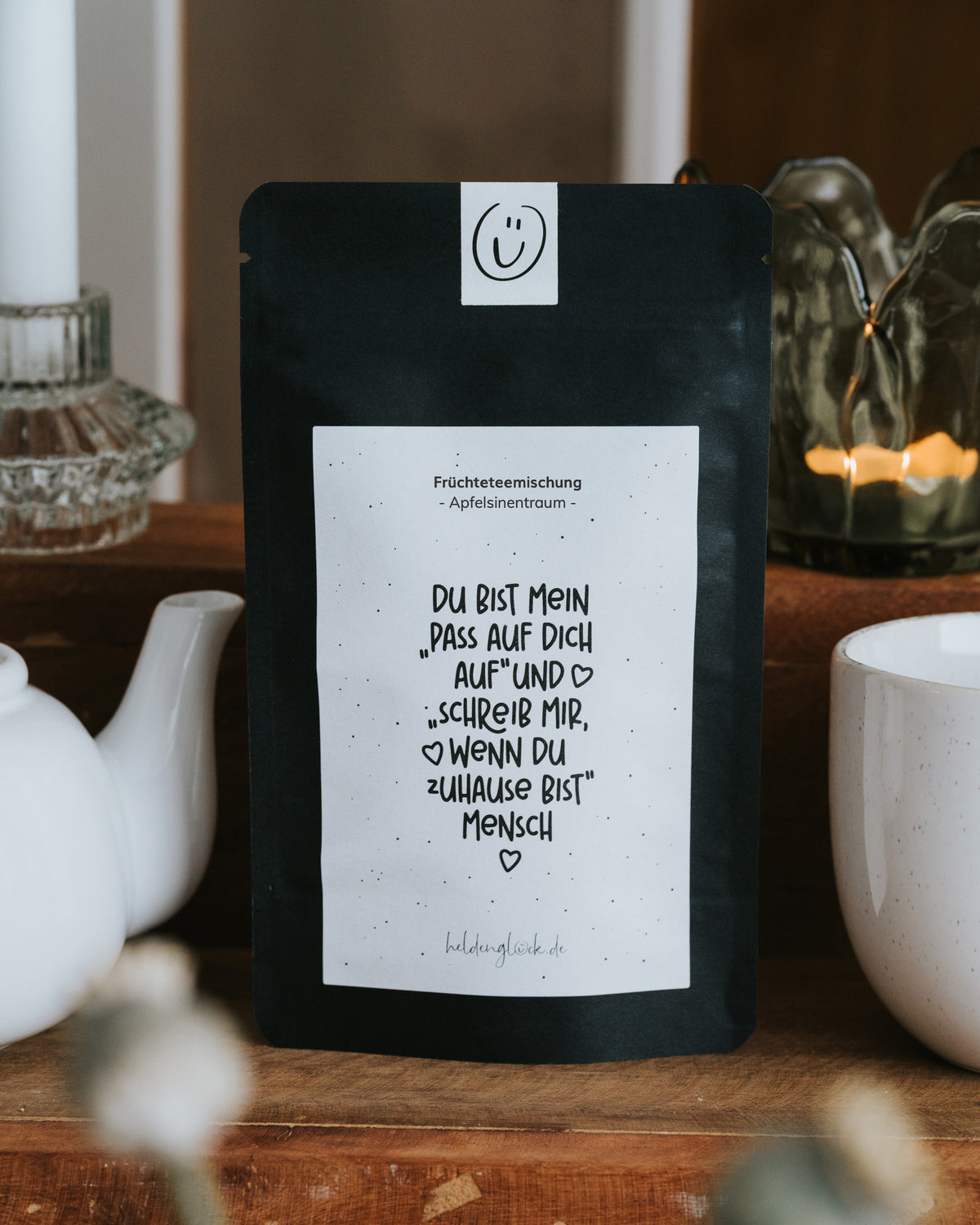 Früchtetee „Pass auf dich auf“ von Heldenglück in schwarzer Aromaschutzverpackung, stehend auf einer Holzkommode zwischen weißer Teekanne und Tasse