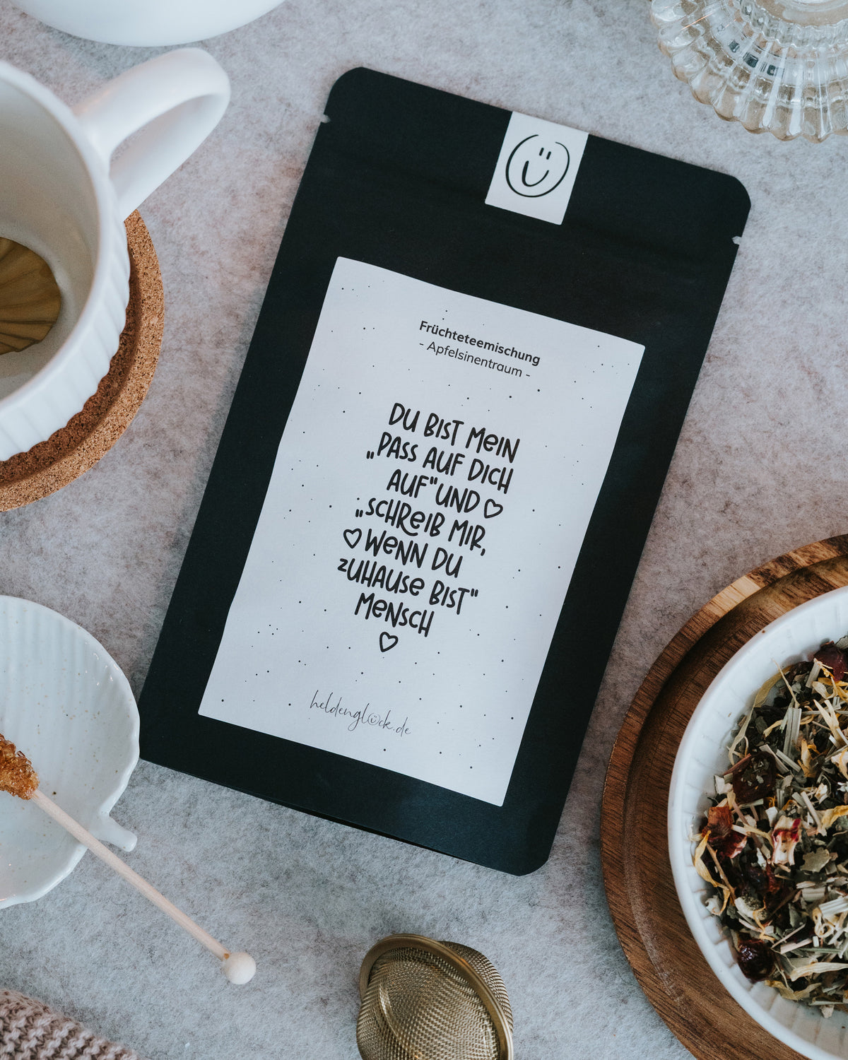 Verpackung der Apfelsinentraum-Früchteteemischung als Flatlay neben aufgebrühter Tasse Tee, goldenem Teesieb und loser Teemischung in kleiner Holzschale