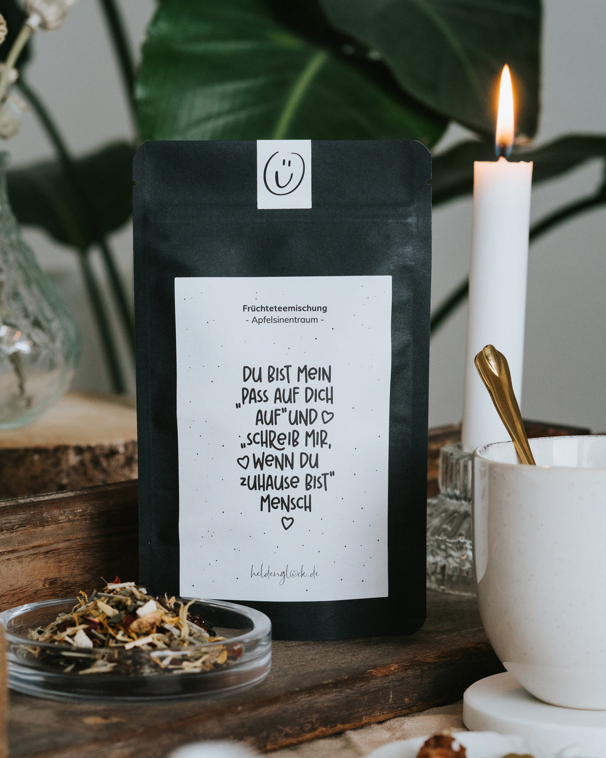 Früchtetee-Beutel mit dem Spruch „Du bist mein Pass-auf-dich-auf-Mensch“ vor grünen Blättern, daneben eine brennende Kerze und eine Tasse mit goldenem Löffel