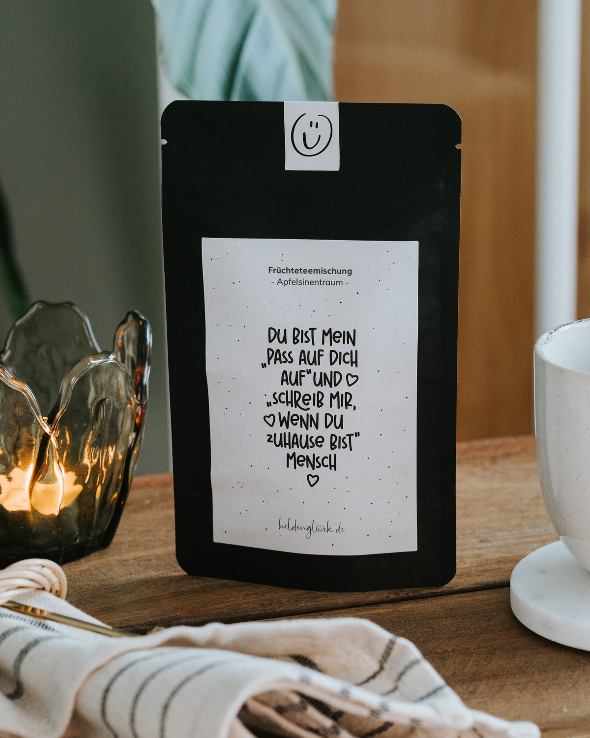 Früchtetee-Verpackung „Pass auf dich auf“ vor einer Zimmerpflanze, mit gläsernem Teelichthalter und heller Tasse als stimmungsvolles Geschenkbild für Freundin oder Mama